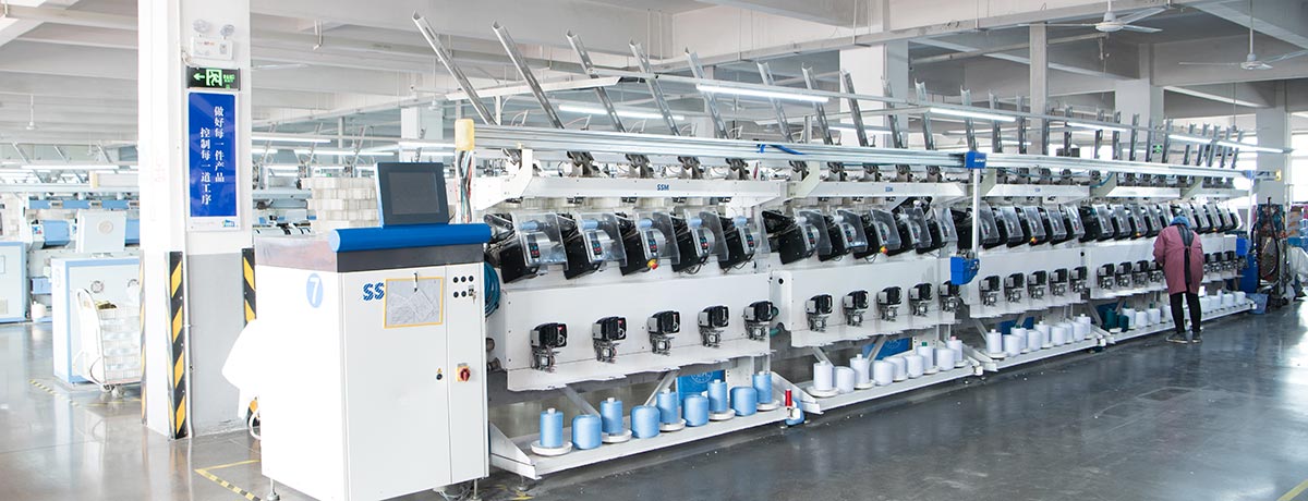 En la imagen se pueden ver trabajadores en una fábrica textil, operando maquinaria de hilatura industrial, mientras manipulan bobinas de hilos de diferentes colores y preparan productos para su distribución en un carrito.